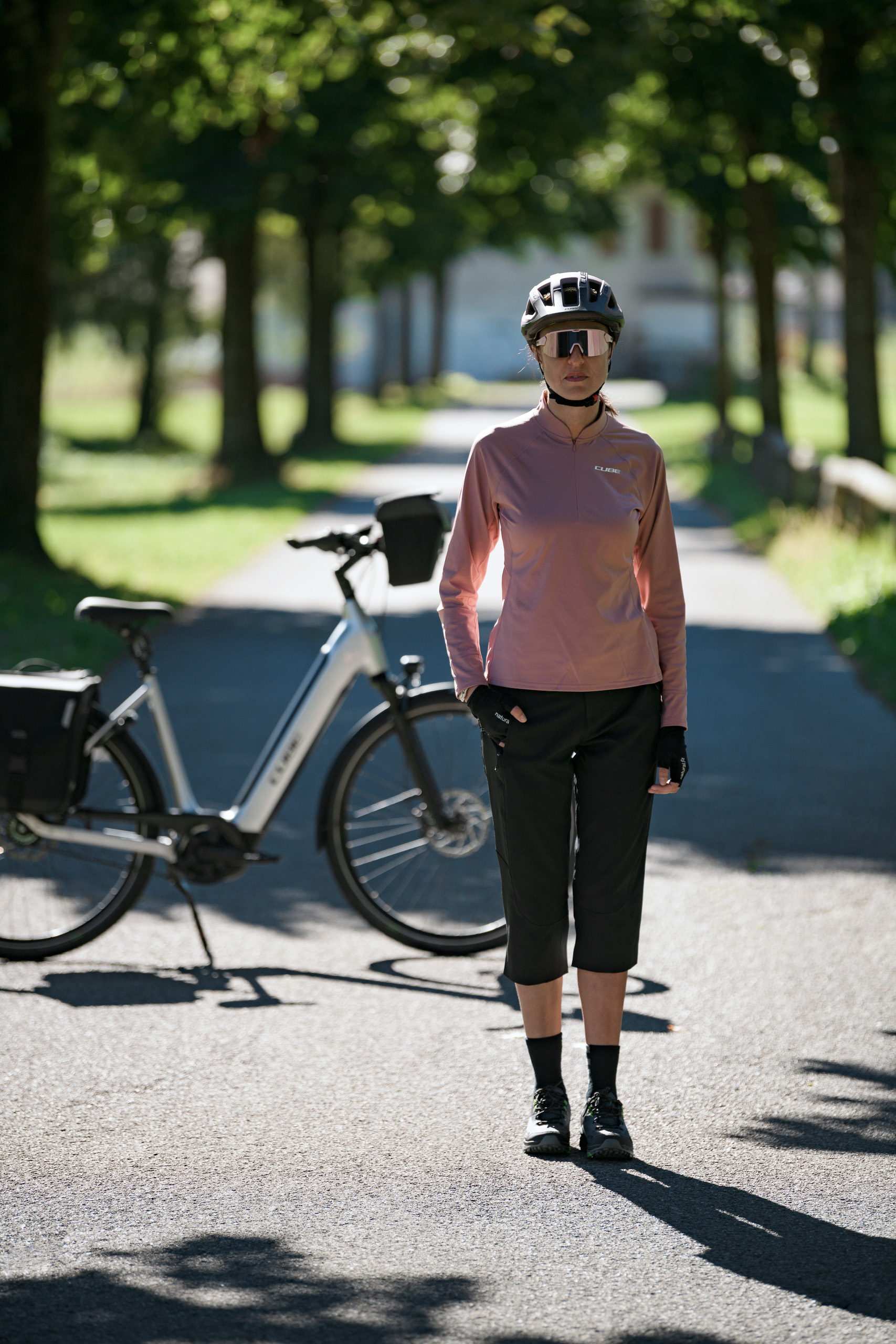 Eine Person mit Helm, Sonnenbrille und Radsportbekleidung steht auf einem sonnenbeschienenen Weg. Ihr Fahrrad ist hinter ihr geparkt. Bäume säumen den Weg und werfen Schatten auf den Boden.