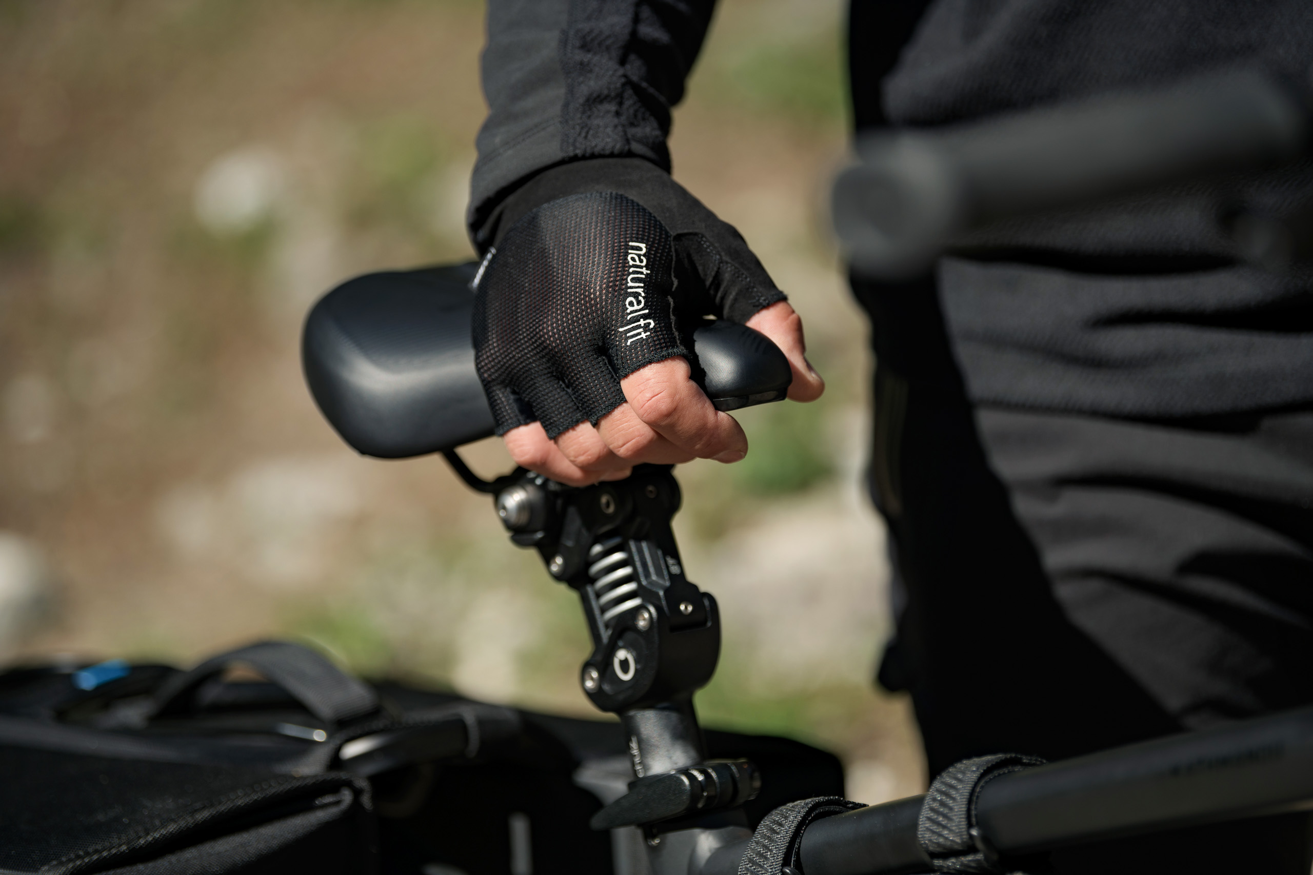Eine Person mit schwarzen Fahrradhandschuhen stellt im Freien den Sattel eines CUBE-Fahrrads ein. Der Fokus liegt auf der behandschuhten Hand und dem Fahrradsitz, im Hintergrund ist unscharfes Grün in der Nähe des CUBE Stores Deggendorf zu sehen.