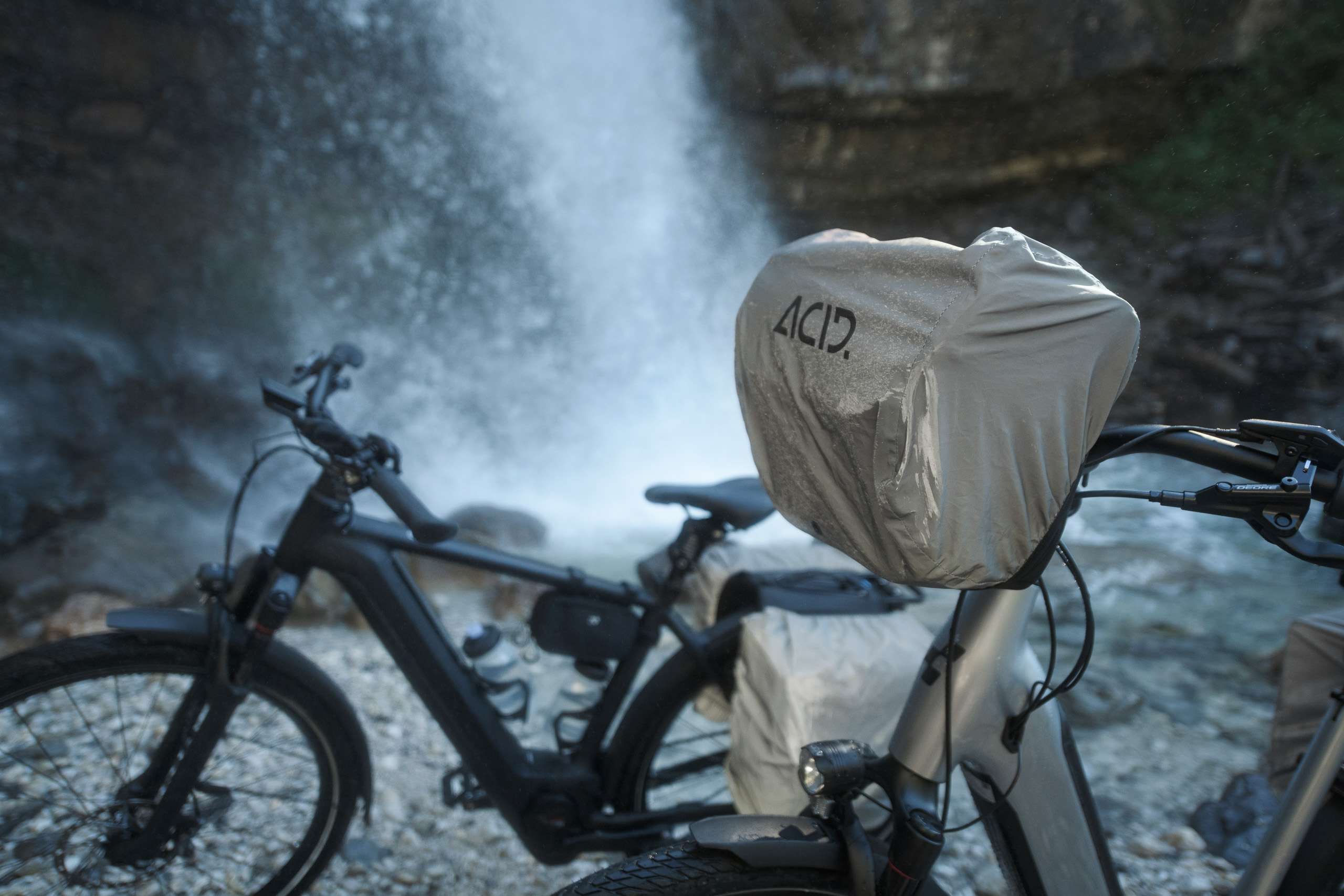 Zwei schwarze CUBE Fahrräder stehen auf felsigem Untergrund in der Nähe eines Wasserfalls in Deggendorf. Sie tragen ACID-Schutzhüllen auf ihren Sitzen und Taschen, im Hintergrund steigt Nebel vom herabstürzenden Wasser auf.