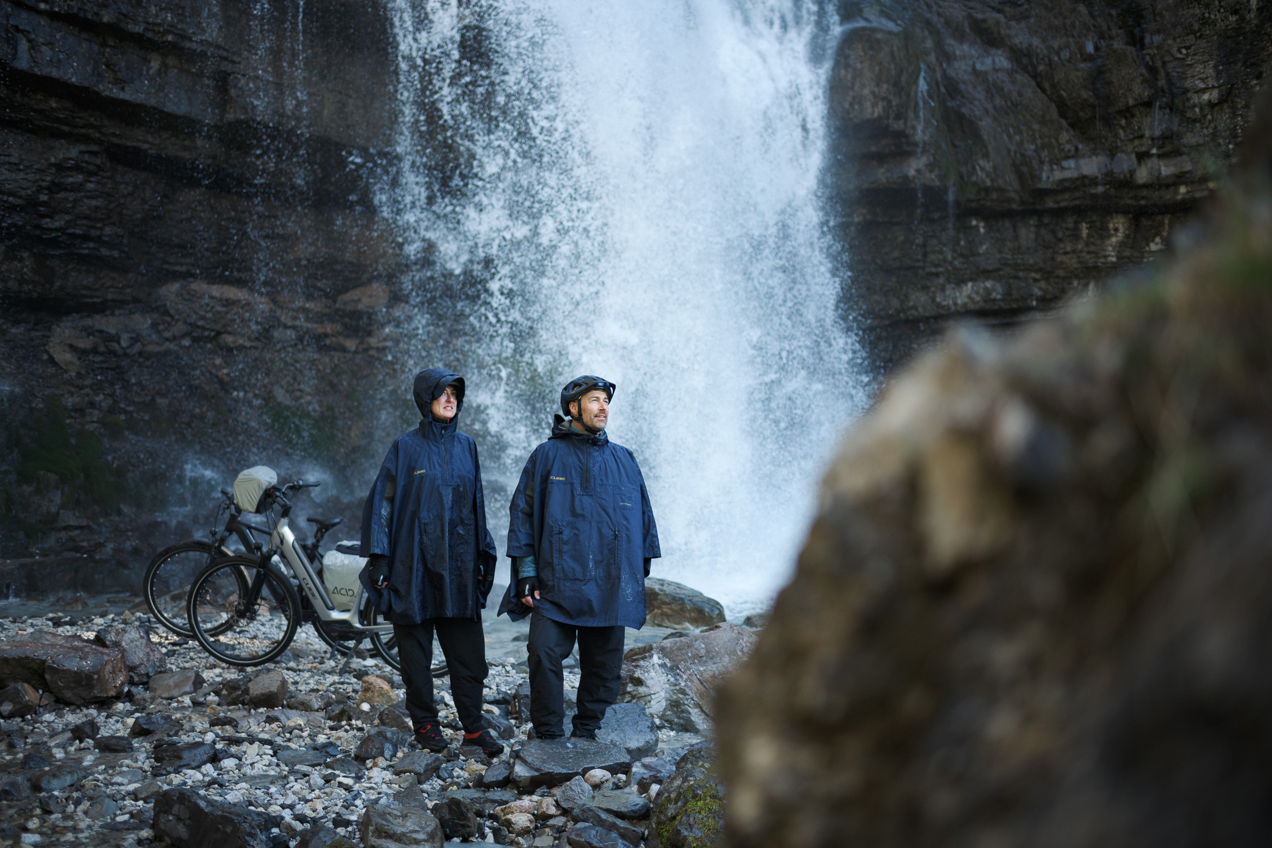 Zwei Personen in blauen Regenponchos und Helmen stehen auf felsigem Boden neben CUBE-Fahrrädern vor einem großen Wasserfall, umgeben von Nebel und schroffen Felsen in der Nähe von Deggendorf.