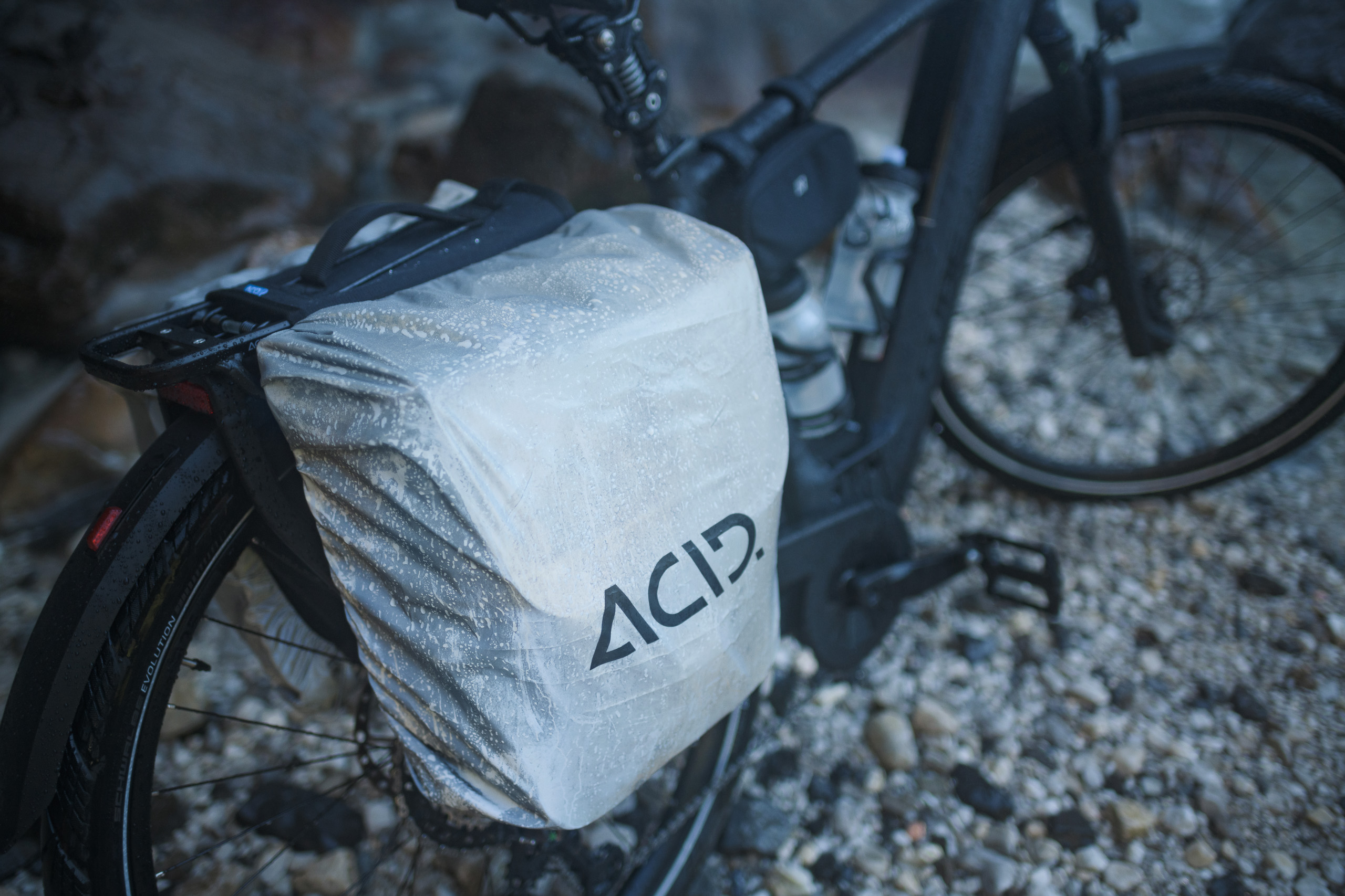 Eine Nahaufnahme eines schwarzen CUBE-Fahrrads mit einer abgedeckten Gepäckträgertasche mit der Aufschrift „ACID“, das auf nassem Schotter im CUBE Store Deggendorf geparkt ist. Wassertropfen perlen auf der durchsichtigen Regenhülle der Tasche.