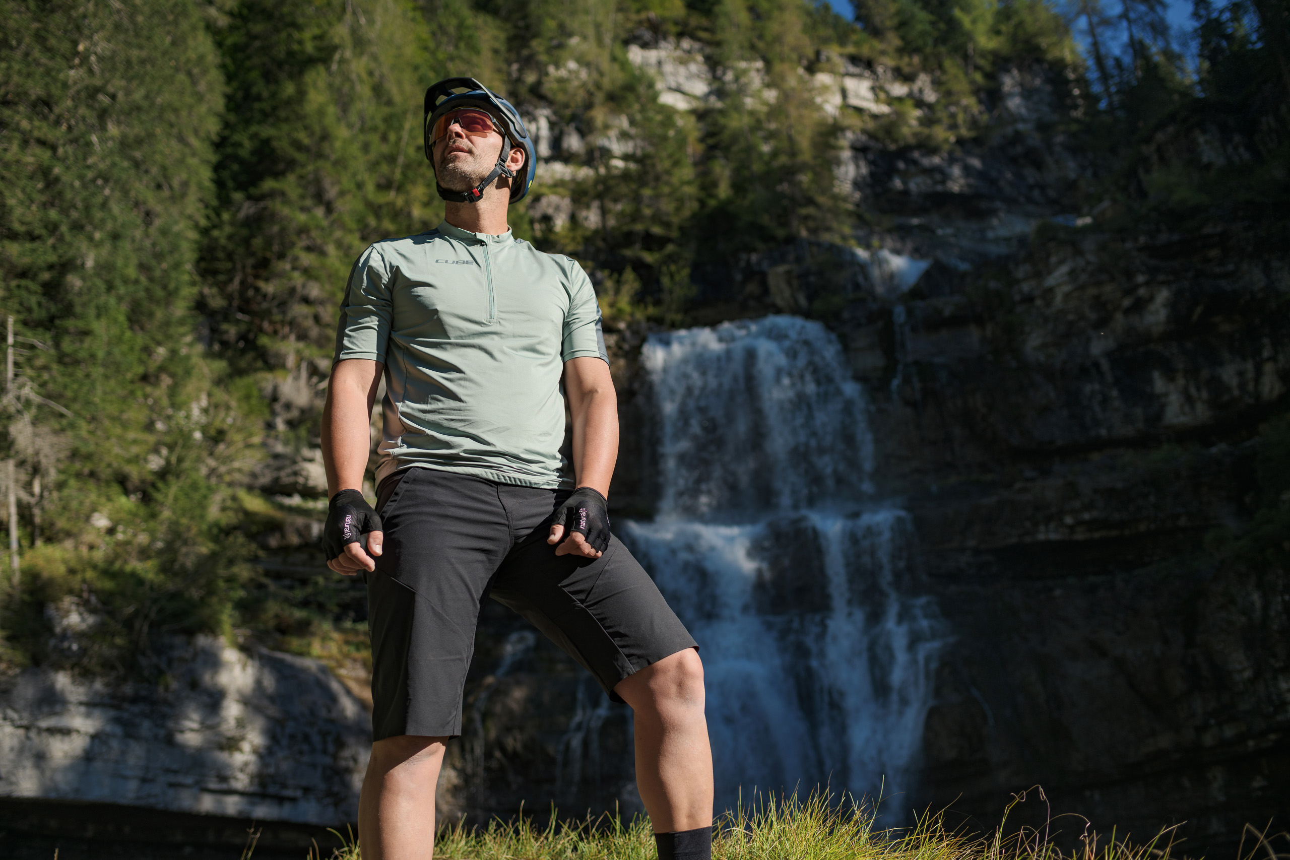 Ein Mann in Radlermontur und Helm steht selbstbewusst mit seinem CUBE Fahrrad auf einer Wiese vor einem Wasserfall bei Deggendorf, umrahmt von bewaldeten Felsen und Sonnenlicht.