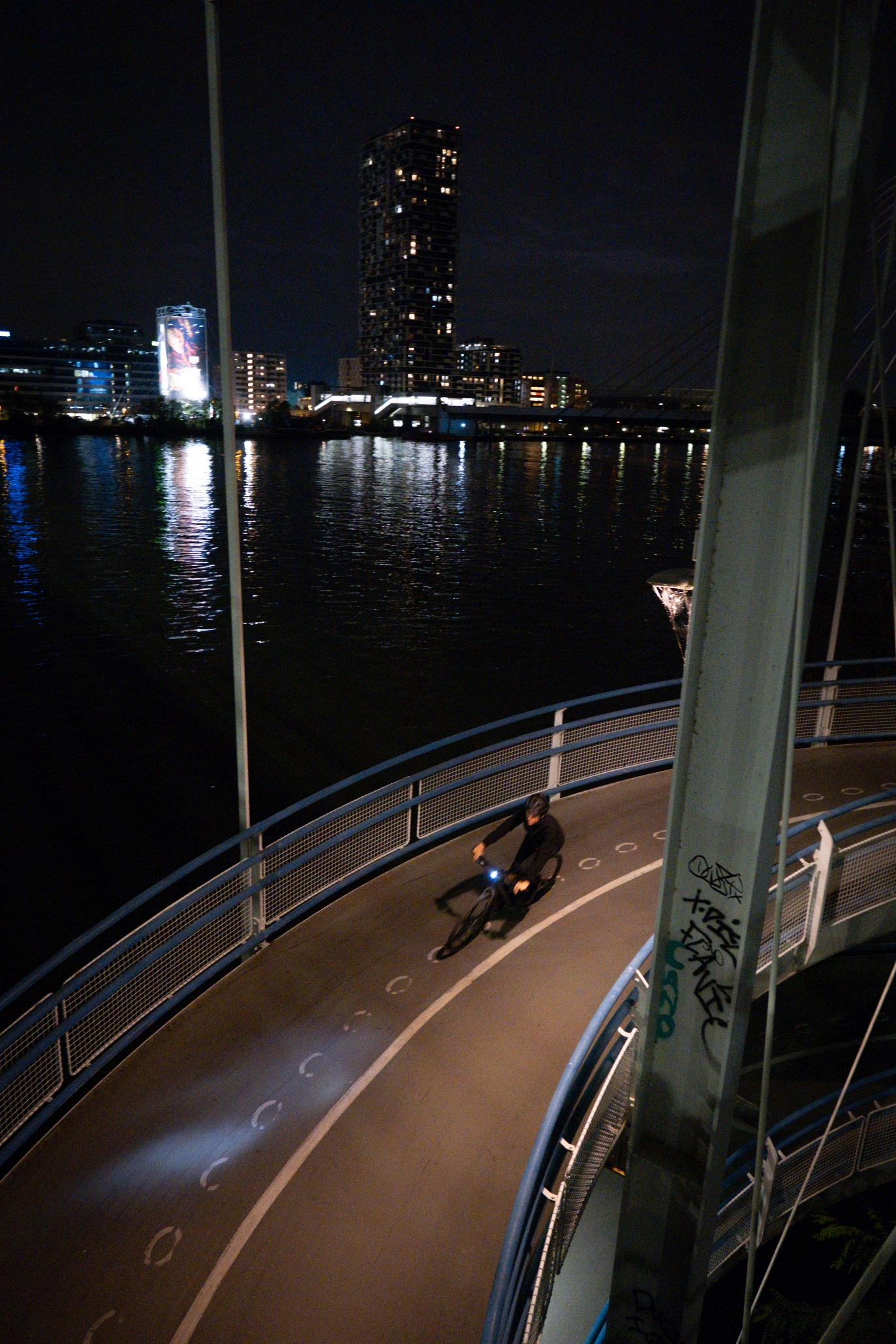 Ein Radfahrer fährt nachts mit einem CUBE Fahrrad auf einem geschwungenen, erhöhten Weg entlang eines Flusses. Im Wasser spiegeln sich die Lichter der Stadt, im Hintergrund sind hohe Gebäude zu sehen. Im Vordergrund sind Graffiti auf einem Pfosten zu sehen.
