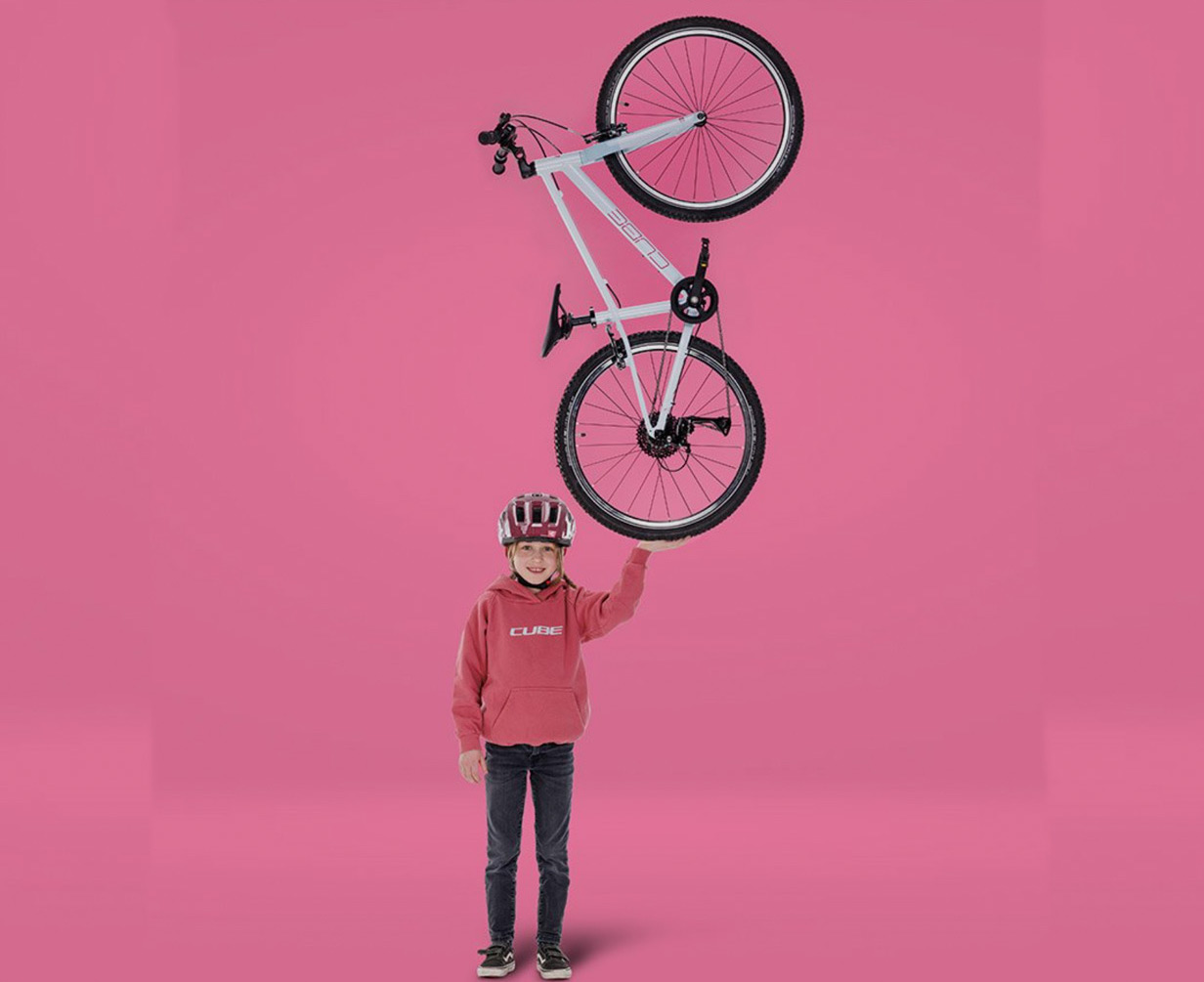 Ein lächelndes Kind mit Helm und rosa Kapuzenpullover balanciert ein CUBE-Fahrrad aufrecht auf einer Hand vor einem einfarbig rosa Hintergrund und ist bereit für das Abenteuer – genau wie im CUBE Store Deggendorf.