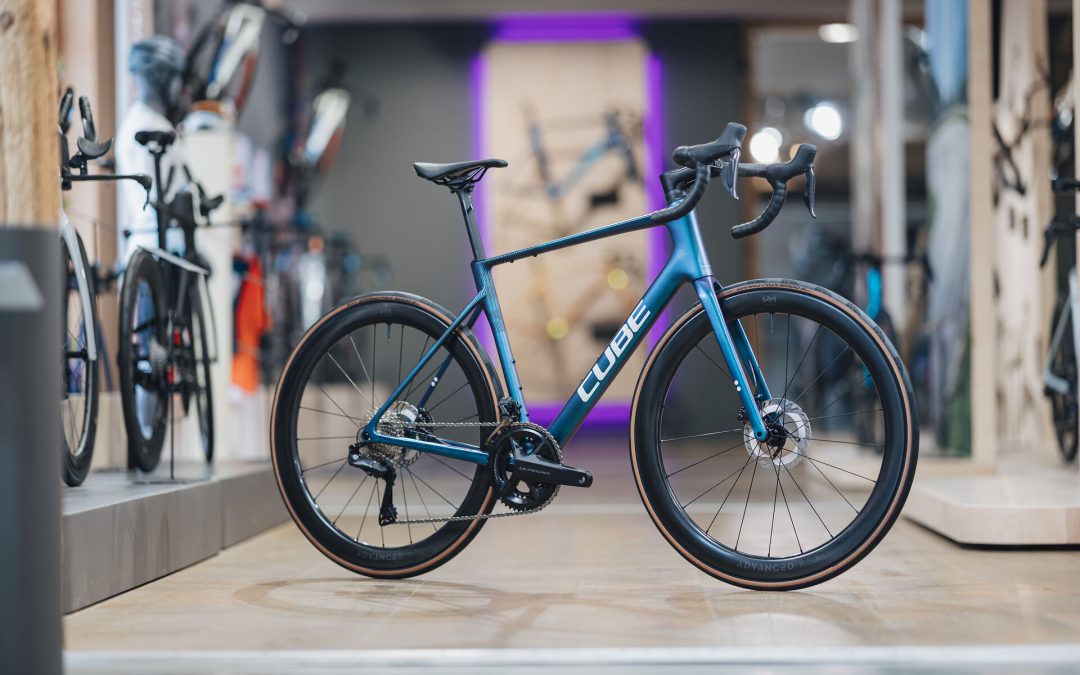 Ein Cube-Rennrad mit blauem und violettem Farbverlauf und abnehmbarem Lenker steht in einem gut beleuchteten Fahrradgeschäft, umgeben von anderen Fahrrädern und Zubehör.